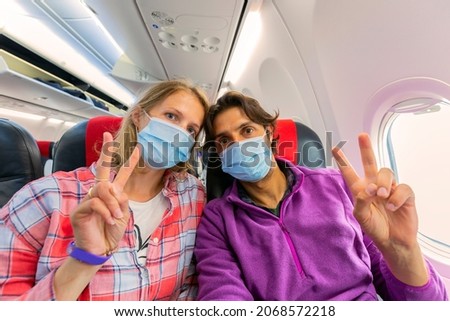 young couple wearing a face mask for COVID-19 protection sitting in an airplane window seat showing two finger V-shaped sign on left hand to the camera during flight - concept