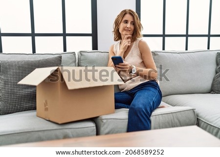 Middle age woman sitting on the sofa checking delivery package on the phone serious face thinking about question with hand on chin, thoughtful about confusing idea 