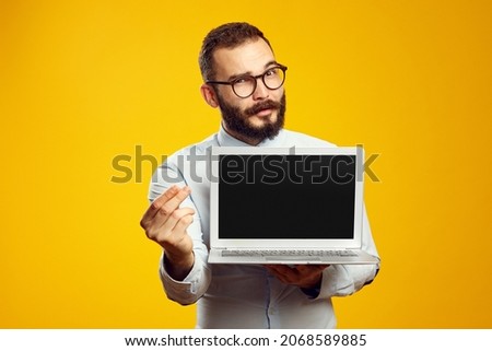 Bearded man in eyeglasses and blue holding new modern laptop device while showing money gesture, isolated over yellow background 