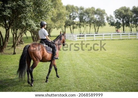 Female horseman riding brown Thoroughbred horse on green meadow in countryside. Concept of rural resting and leisure. Green tourism. Young smiling european woman. Beautiful landscape at sunny day Royalty-Free Stock Photo #2068589999