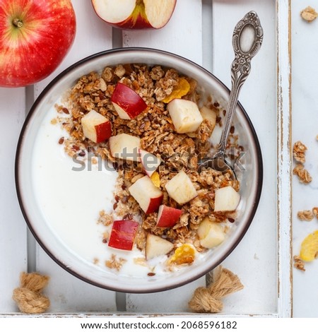 Homemade granola (muesli) with oatmeal, nuts, flaxseed, cornflakes, fresh red apple, cinnamon and yogurt. Delicious healthy breakfast on a white plate on a marble background. Diet. Proper nutrition 