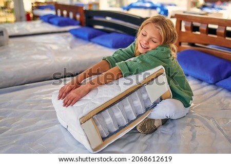 Beautiful child cute girl sitting on the bed and holding a cutaway mattress sample in the store. Purchase of bedding. Royalty-Free Stock Photo #2068612619