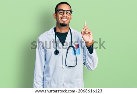 Young african american man wearing doctor uniform and stethoscope showing and pointing up with finger number one while smiling confident and happy. 