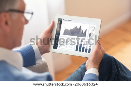 Serious elderly caucasian man in glasses works at tablet with charts on screen, at home interior, over shoulder view. Passive income, successful retirement business, finance app and profit indoor