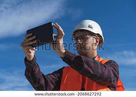 Adult man working in a wind farm using his tablet to take a photo. He is wearing a safety helmet and a reflective vest.