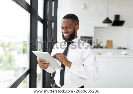 Smiling African American man using computer tablet, standing in modern coworking office, looking at screen, confident young male checking email, working online, writing message, browsing internet
