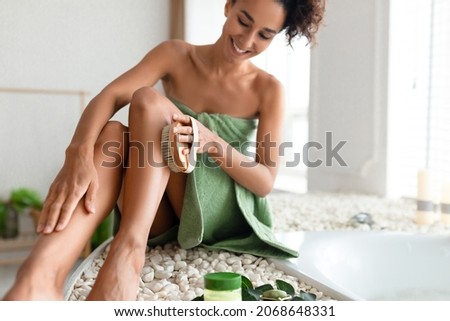 Young woman in towel making anti cellulite or lymphatic drainage massage near foamy bath at home, selective focus. Millennial lady dry brushing her leg, exfoliating skin, pampering herself in bathroom Royalty-Free Stock Photo #2068648331
