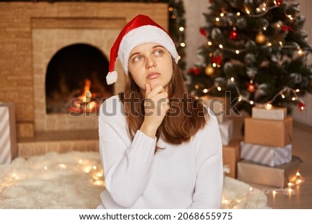 Portrait of pensive woman wearing white sweater and santa claus hat, sitting near fireplace and xmas tree in festive living room, holding chin, thinking who invite to celebrate Christmas.