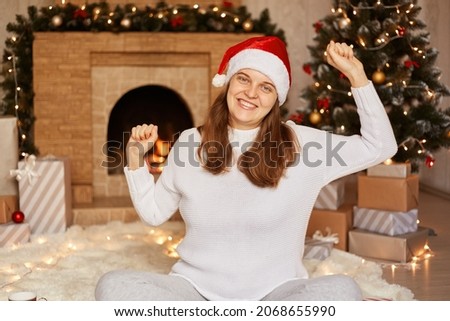 Positive young adult beautiful woman wearing white sweater and santa claus hat, posing near fireplace and xmas tree, sitting with raised arms, dancing happily, celebrating New year eve.