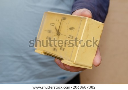 A man's hand holds out an old table clock to the camera. A rectangular alarm clock in a yellowed plastic case. Indoors. Selective focus.