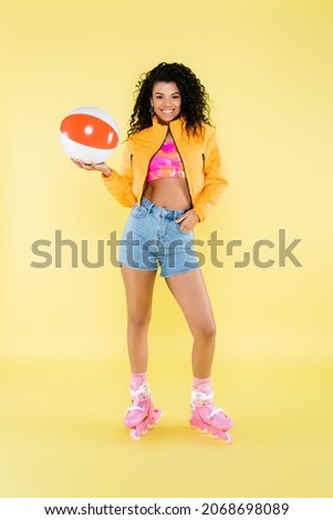 full length of positive african american young woman on roller skates holding inflatable ball on yellow