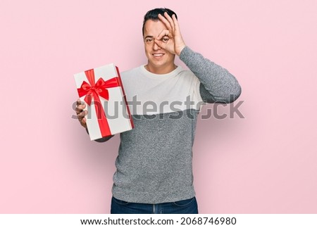 Handsome young man holding gift smiling happy doing ok sign with hand on eye looking through fingers 