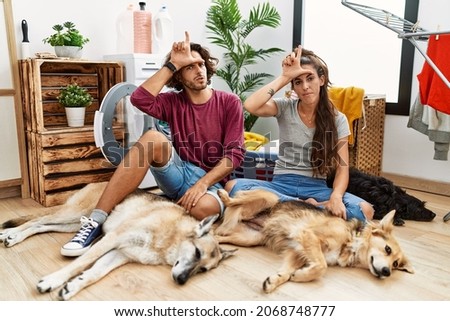 Young hispanic couple doing laundry with dogs making fun of people with fingers on forehead doing loser gesture mocking and insulting. 