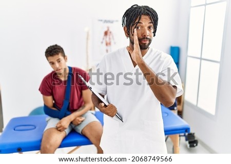 Young hispanic man working at pain recovery clinic with a man with broken arm hand on mouth telling secret rumor, whispering malicious talk conversation  Royalty-Free Stock Photo #2068749566