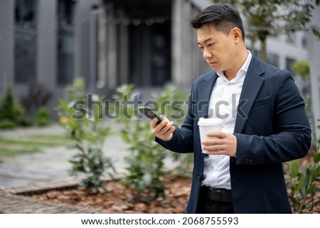 Asian businessman with coffee cup calling on smartphone on city street. Concept of modern successful man. Idea of remote and freelance work. Person wearing suit