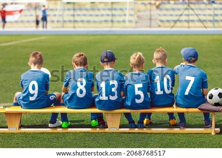School Boys in Sports Football Team. Kids is Classic Blue Soccer Jersey Uniforms With Numbers. Kids Playing Sports Together. Children Sitting on Sideline Wooden Substitiute Bench Royalty-Free Stock Photo #2068768517