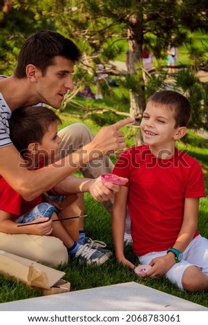 dad plays and fools around with his sons while drawing a picture, getting dirty in paint and laughing cheerfully in the open air in the park on a sunny warm summer day