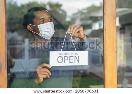 African american business man with face mask holding the sign for the reopening and looking away of the place after lockdown quarantine. Small business owner reopen and back to new normal concept.
