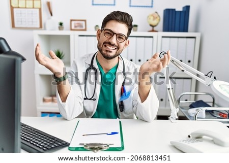 Young man with beard wearing doctor uniform and stethoscope at the clinic smiling cheerful offering hands giving assistance and acceptance. 
