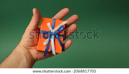Man's hand holding a red gift box with white and blue bow on green paper background.