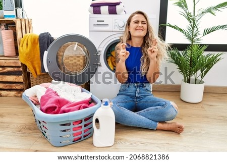 Young beautiful woman doing laundry sitting by wicker basket gesturing finger crossed smiling with hope and eyes closed. luck and superstitious concept. 