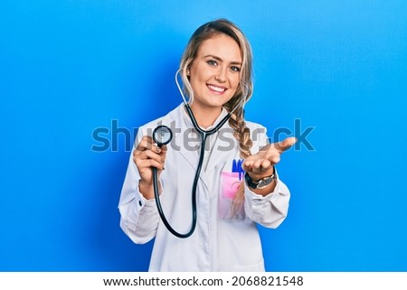 Beautiful young blonde doctor woman holding stethoscope smiling cheerful offering palm hand giving assistance and acceptance. 