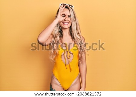 Beautiful young blonde woman wearing swimsuit and sunglasses doing ok gesture with hand smiling, eye looking through fingers with happy face. 