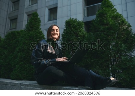 Lifestyle portrait of a young cute woman freelancer, copywriter, motivated start-up sitting on parapet with laptop, working remotely and smiling cute for camera. Connection and online business concept