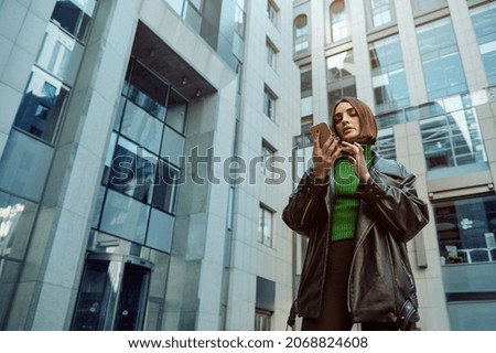 Beautiful self-confident European young woman, freelancer, entrepreneur, copywriter using mobile phone on modern office corporate high-rise buildings background. Business and communications concept.