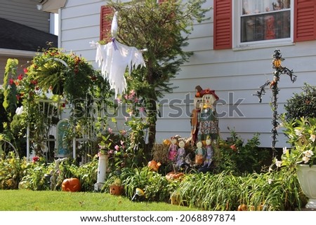 A festive fall display in the flower bed of a suburban home. There are three bales of hay, a flag that reads "Happy Fall Y'All", pumpkins, and decorations in the ground.
