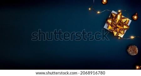 Christmas gift. White gift with golden bow, gold balls and sparkling lights garland in xmas decoration on dark background for greeting card. Xmas backdrop with space for text