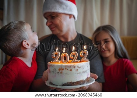 Dad with two children looks dreamily at burning candles on Christmas cake with golden 2022 numbers on top with yellow icing over bokeh lights background, family New Year celebration, focus on candles