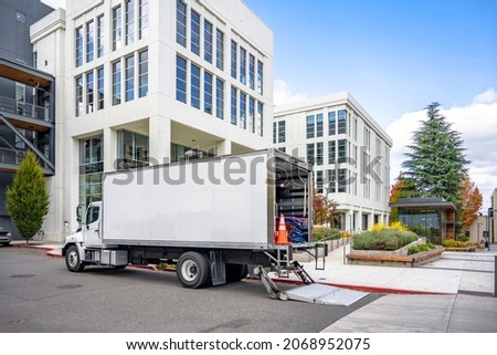 White middle class relocation rig day cab semi truck tractor with long spacious box trailer standing on the urban city street with multilevel apartment and office buildings unloading delivered goods Royalty-Free Stock Photo #2068952075