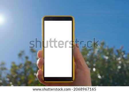 Hand holding a smartphone vertically, outdoors, with sky and greenery in the background, a sunny day