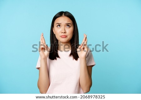 Worried asian girl looking up hopeful and nervous, cross fingers for good luck, making wish, praying, standing in t-shirt over blue background