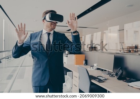 Young man in formal suit enjoying virtual tour or excursion with VR headset. Businessman in virtual reality glasses standing near working desk in office, tries to touch objects in digital actuality Royalty-Free Stock Photo #2069009300