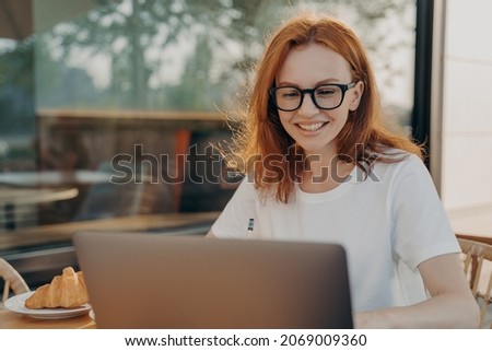 Beautiful redhead professional woman freelancer works on laptop computer poses in outdoor cafe wears casual white t shirt spectacles chats in social networks has morning breakfast in coffee shop Royalty-Free Stock Photo #2069009360