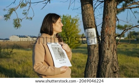 woman with flyers stack stands near poster report of missing dog with photo stuck on tree trunk closeup