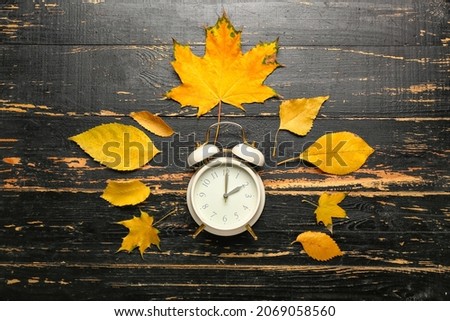 Alarm clock and autumn leaves on black wooden background. Daylight saving time end
