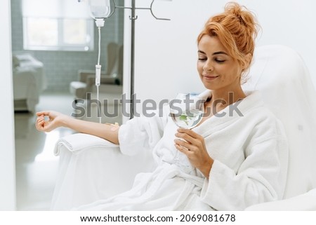 Waist up portrait view of the charming woman in white bathrobe sitting in armchair and receiving IV infusion. She is holding glass of lemon beverage and smiling  Royalty-Free Stock Photo #2069081678