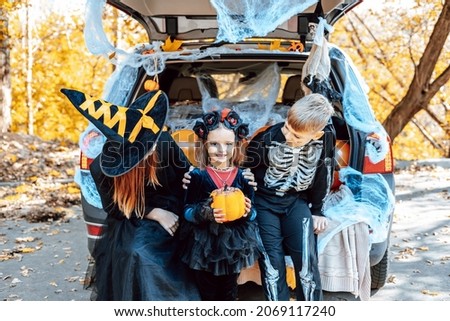 siblings boy in skeleton costume, teenage girl in witch costume and hat and cute little girl in spooky costume sits in trunk car decorated for Halloween with web, orange balloons and pumpkins, outdoor