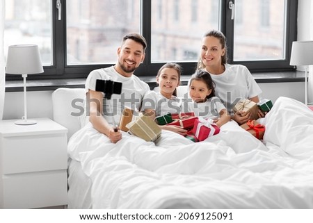 family, winter holidays and people concept - happy mother, father and two daughters with christmas gifts taking selfie on smartphone in bed at home