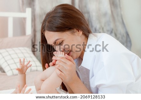 Young  mother kissing boy toddler son feet heels legs. Mom playing with child baby on bed in bedroom at home. Happy authentic family childhood lifestyle. Tender touching sweet moments.