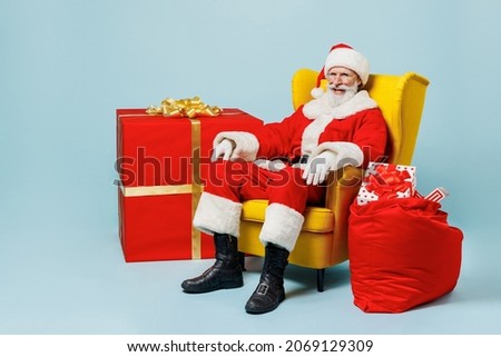 Full body fun old Santa Claus man 50s in Christmas hat red suit sit in armchair near gifts bag isolated on plain blue background studio. Happy New Year 2022 celebration merry ho x-mas holiday concept