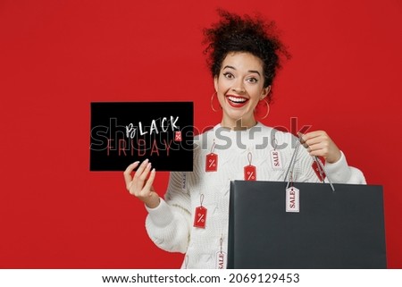 Young fun happy woman in white sweater with tags sale in store showroom hold package bags with purchases after shopping card sign black friday text inscription isolated on plain red background studio