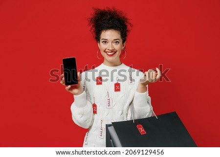 Young fun happy woman in white sweater with tags sale hold package bags with purchases after shopping using mobile cell phone with blank screen workspace area isolated on plain red background studio.