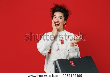 Young happy amazed female costumer woman wear white knitted sweater with tags sale in store showroom hold package bags with purchases after shopping cover mouth isolated on plain red background studio