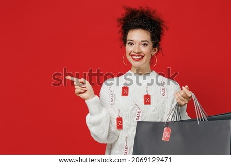 Young happy female costumer woman wear white knitted sweater with tags sale hold package bags with purchases after shopping point finger aside on workspace area isolated on plain red background studio