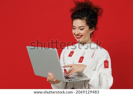 Young smiling happy caucasian female costumer woman 20s wear white knitted sweater with tags sale in store showroom hold use work on laptop pc computer isolated on plain red background studio portrait