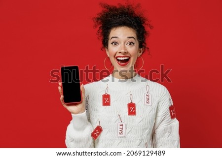 Young female costumer woman 20s wear white knitted sweater with tags sale in store showroom hold in hand use mobile cell phone with blank screen workspace area isolated on plain red background studio